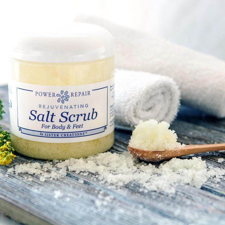 Power Repair Salt Scrub by Sister Creations sitting on a wood board with rolled white towels and flowers surrounding it.