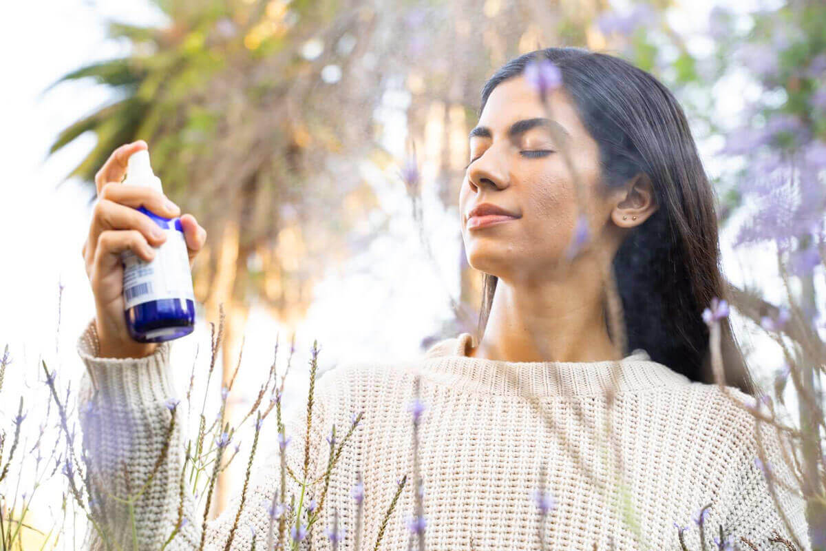 A woman Sprays herself with the Power Repair face mist. She is standing amongst trees and flowers and look quite serene. Edit alt text