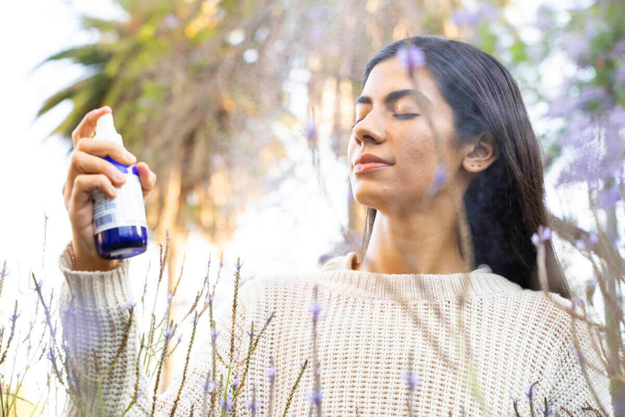 A woman Sprays herself with the Power Repair face mist. She is standing amongst trees and flowers and look quite serene.  Edit alt text