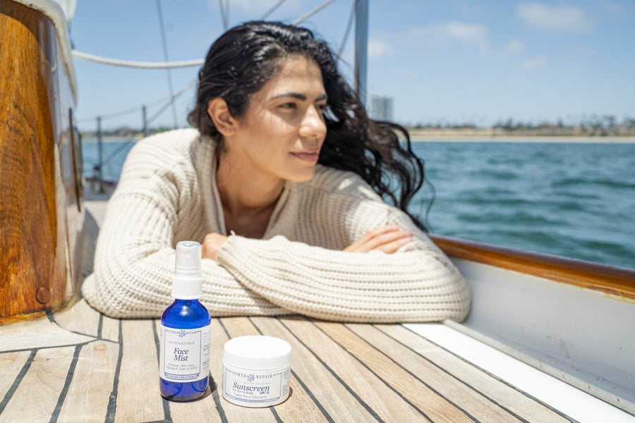 A Pretty woman is lounging on a boat and looking off into the distance across the ocean. The Power Repair Face Mist and Power Repair sunscreen are sitting in the foreground.