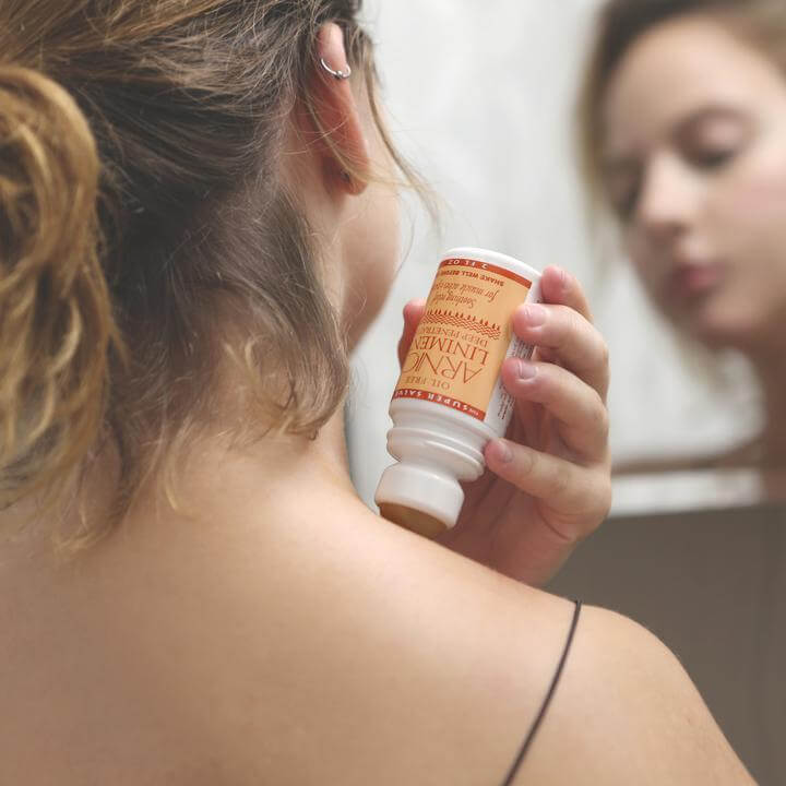 A woman subs the Arnica Linament Roller onto her shoulder, while looking in the mirror.