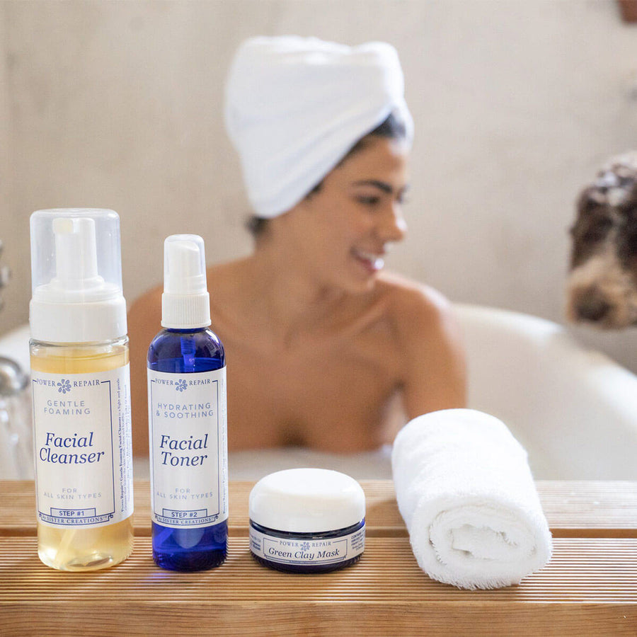A woman is smiling in the bathtub while her cute dog with curly hair peers in. The Power Repair Skin Care line is sitting in front of her on a shelf.