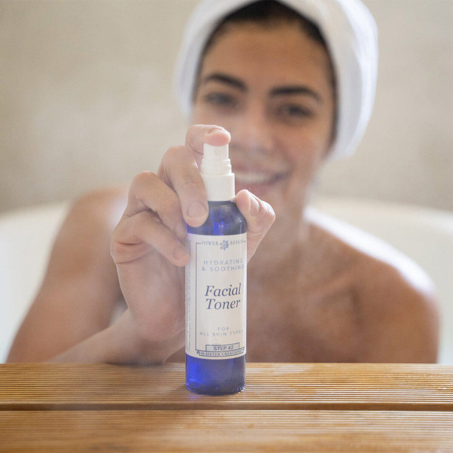 A woman joyfully spraying the Power Repair Facial Toner, while she is smiling in the bath tub.