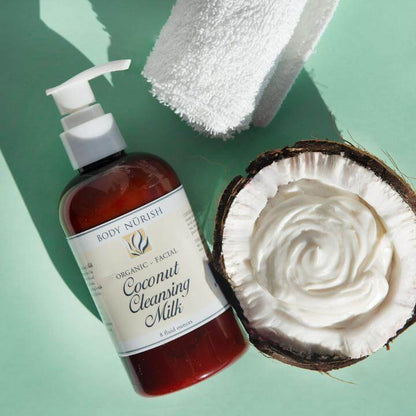 A bottle of coconut cleansing  milk laying on a seafoam colored background.  It is sitting next to a coconut that is cut in a half with the creamy cleansing milk inside.  There is a a white rolled up towel in the frame.