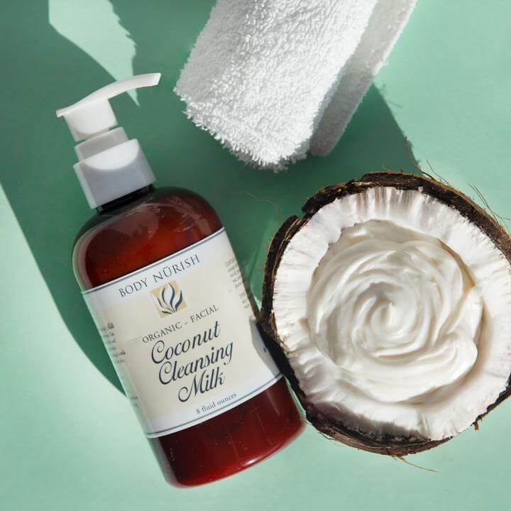 A bottle of coconut cleansing  milk laying on a seafoam colored background.  It is sitting next to a coconut that is cut in a half with the creamy cleansing milk inside.  There is a a white rolled up towel in the frame.