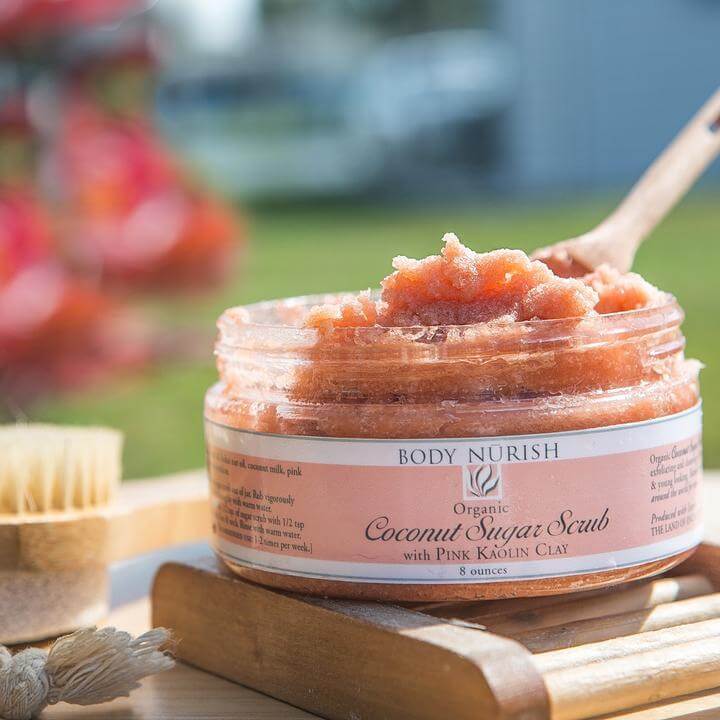 Body Nurish Coconut Sugar Scrub 8oz. sitting on a wooden table with a wooden spoon in it shoeing its luxurious texture, with a peacful outdoor scene in the background