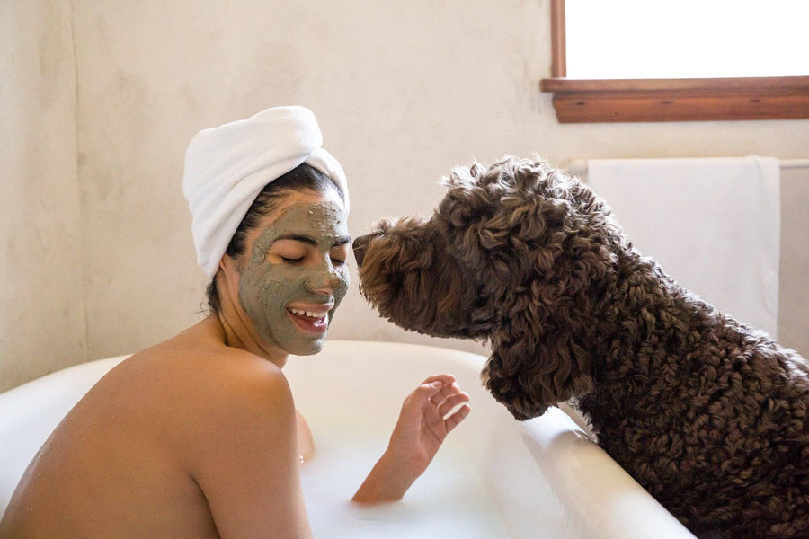 I woman Sits in the bath look relaxed with the Body Nurish Green Clay Refinisher Mask on her face while her cute dog looks in.