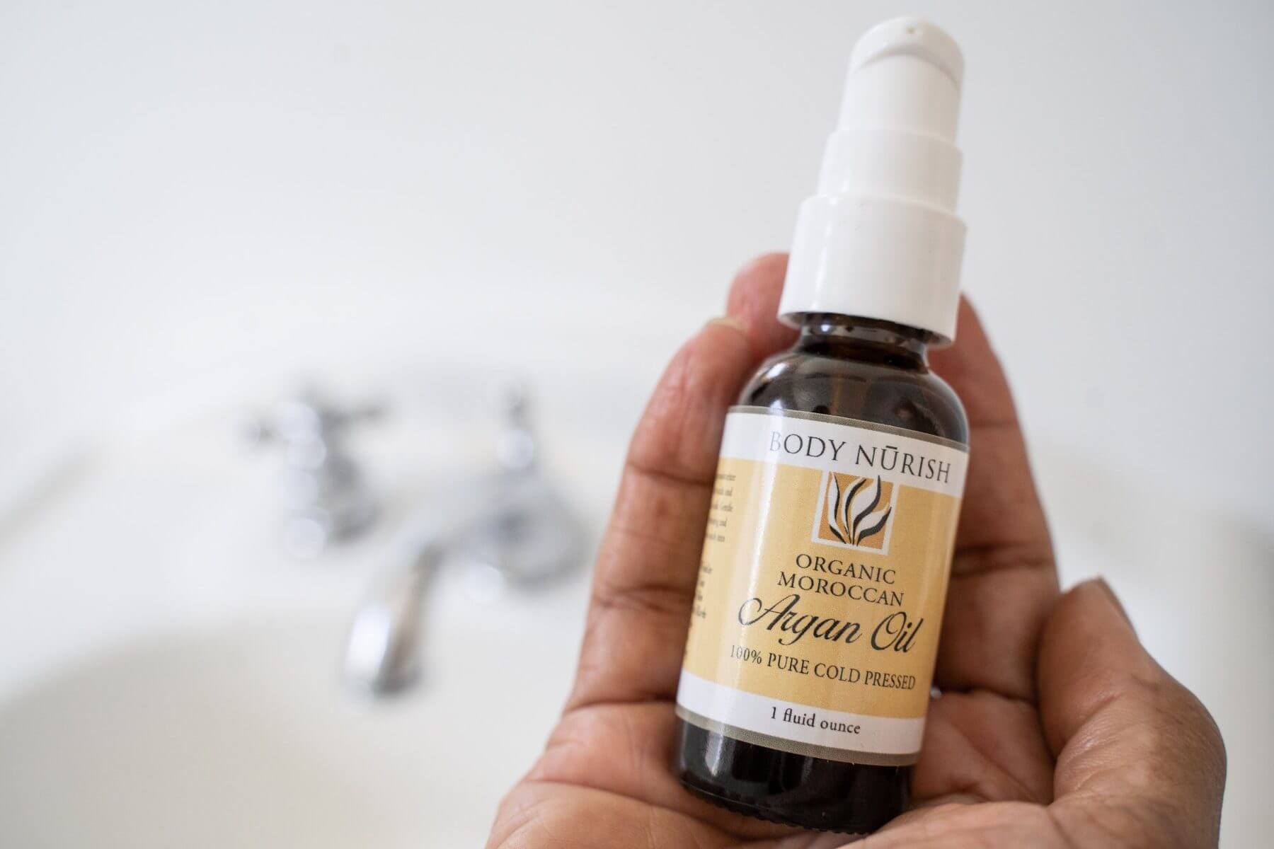 1 oz. Body Nurish Organic Moroccan Argan Oil buin gheld in a hand with a crisp white sink in the background.