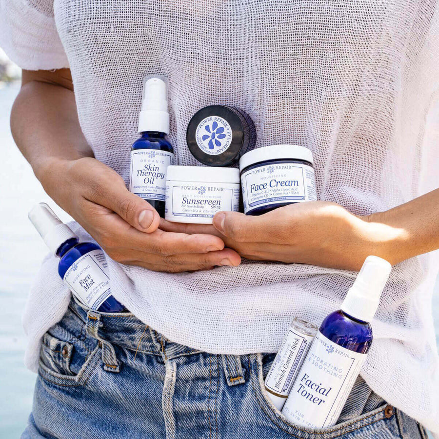A woman at the beach holds an armful of Power Repair products like Power Repair Eye Cream, Power Repair Face Mist, Power Repair Sunscreen, and more Power Repair products bulge from her jean shorts pockets