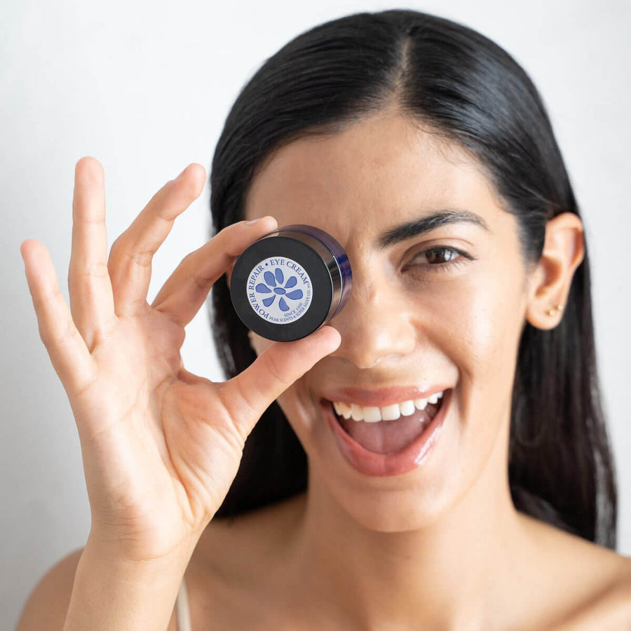 A woman with clear healthy skin smiles and holds a round container of Power Repair eye cream in front of her eye