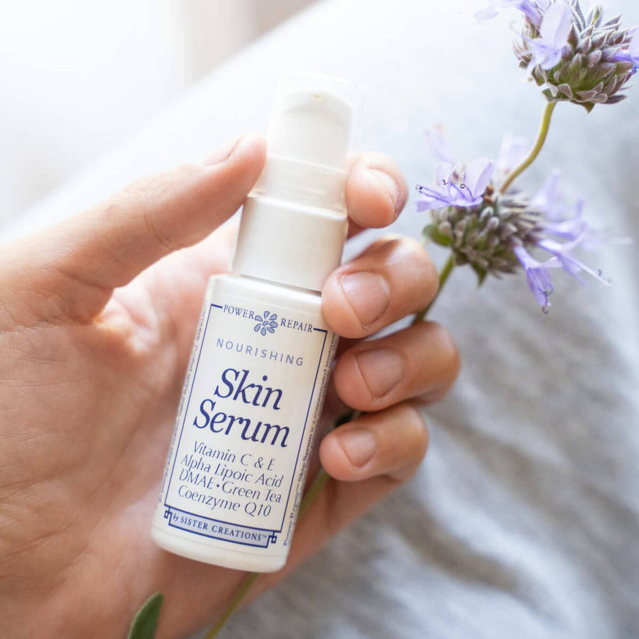 A 1 oz. bottle of Power Repair Skin Serum by Sister Creations rests in the palm of a hand next to a lavender flower, which is an ingredient in Power Repair Skin Serum