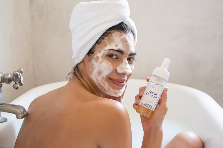 A woman in a bath tub has Power Repair Skin Cleanser by Sister Creations into her face. It is Foamy, she looks happy, and there is a towel on her head.