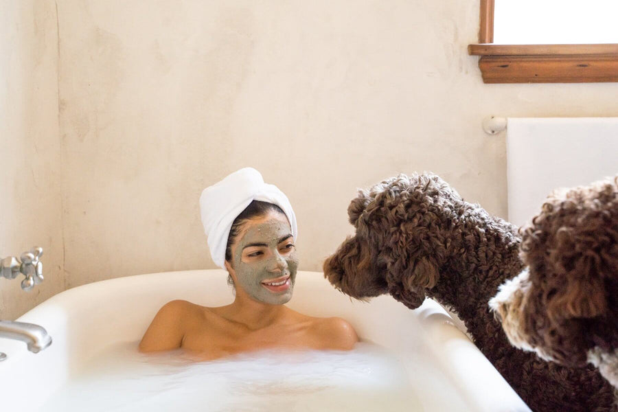 A Woman is wearing Power Repair Green Clay Mask by Sister Creations while she is relaxing in the bath and her two cute, curly haired dogs are looking at her.
