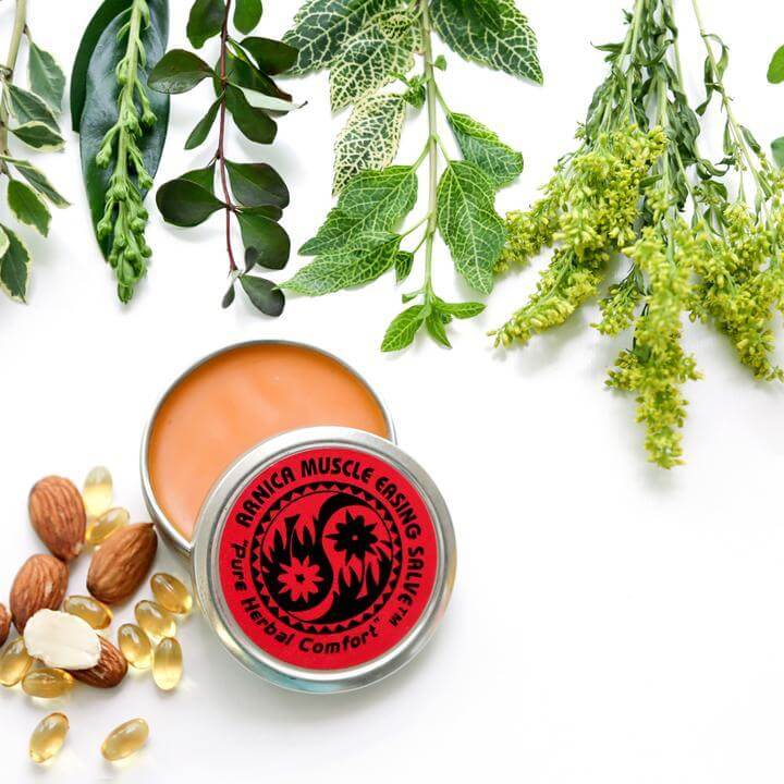 A tin of Arnica Muscle Easing Salve sit open, showing its lovely orange color. There are plants, almonds, and oil capsules beautifully displayed around it.