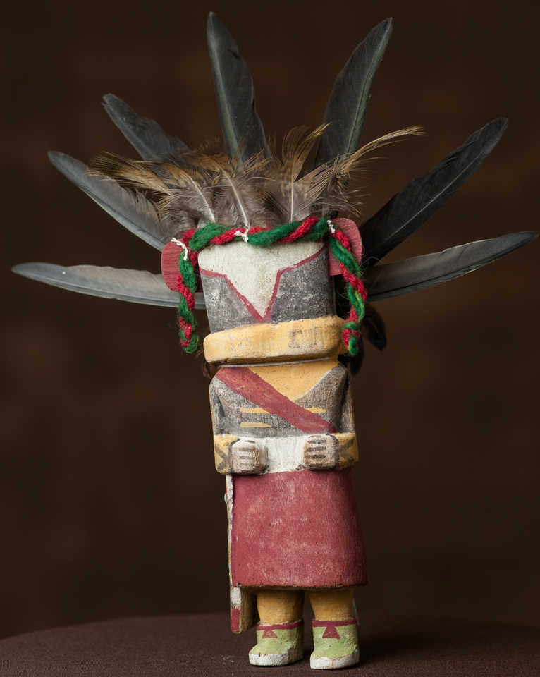 The Blue Chasing Star Katsina doll carved in the traditional style by Philbert Honanie features beautiful, worn-looking black, red, yellow, and white naturally pigmented colors and is adorned with feathers and strands of yarn