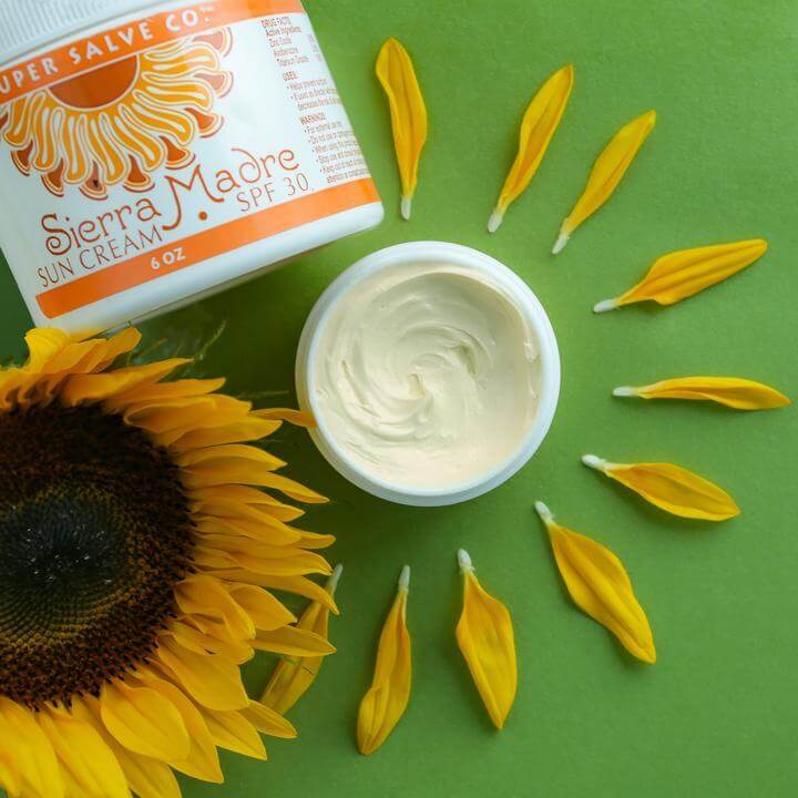 Sierra Madre Sun Cream 6 oz. sitting next to a beautiful sunflower on a green background. An open container of the sierra madre sun cream is sitting next to it with sunflower petals all around it.