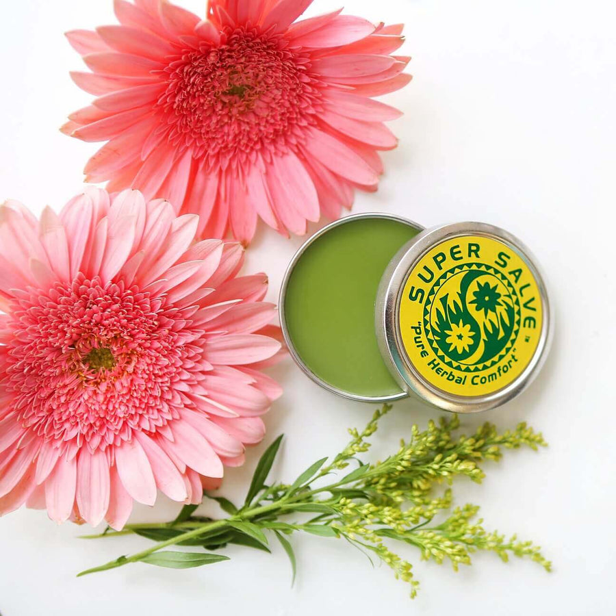 A tin of Super Salve sit with its lid cracked to expose the vibrant green color of the salve. Next to it are beatiful pink and yellow flowers.