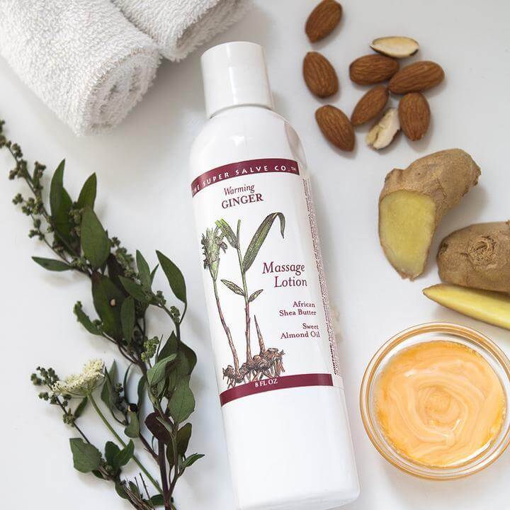 Warming Massage Lotion with Ginger lays on a white background with some of the ingredients in the lotion displayed beautifully around it. There are almonds, ginger, and jojoba leaves.